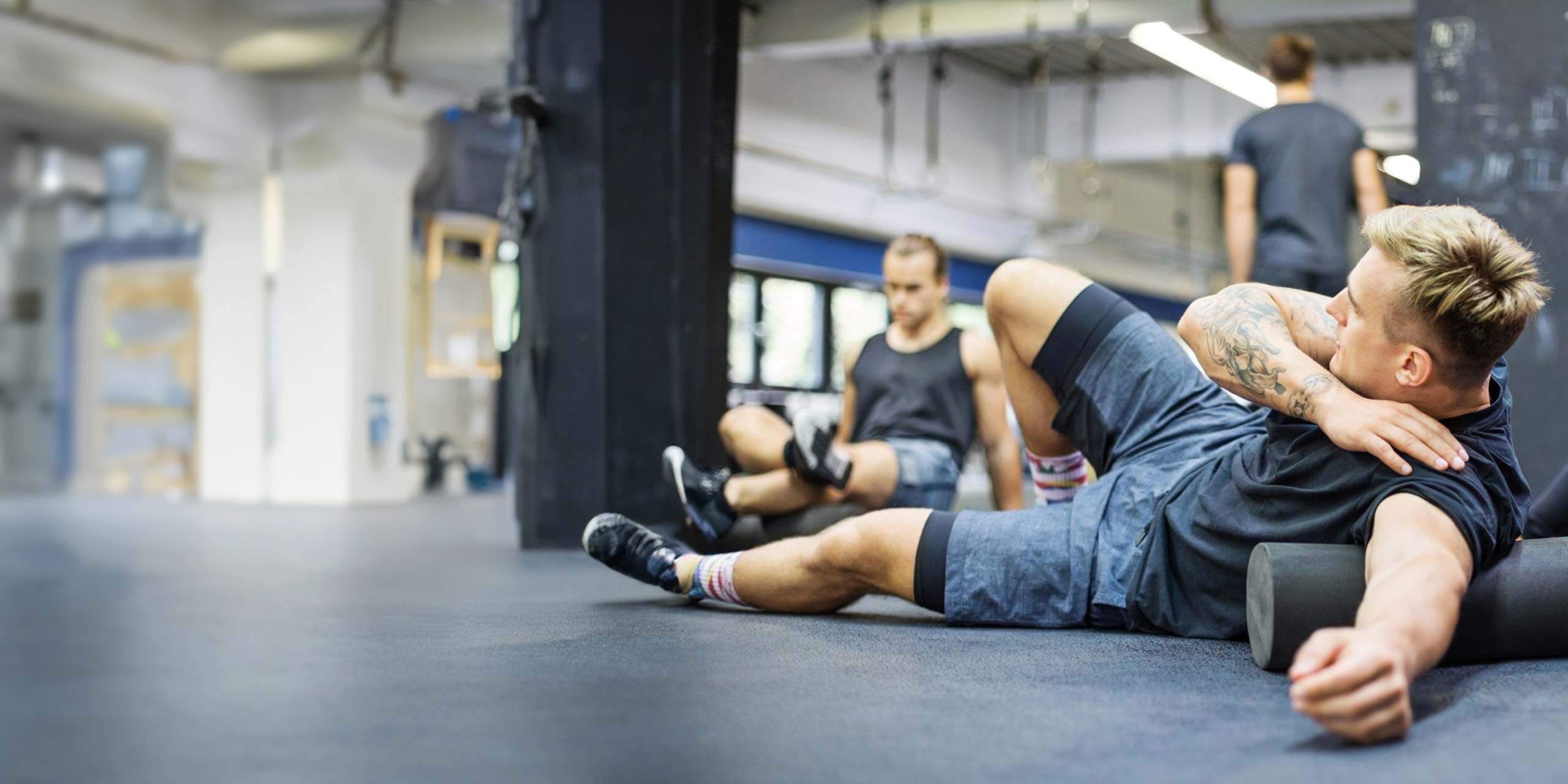 Ein Mann nutzt in einem Fitnessstudio eine Schaumstoffrolle zur Muskelregeneration, während eine andere Person im Hintergrund trainiert und so die Wirksamkeit der Faszienrolle-Anwendung demonstriert.
