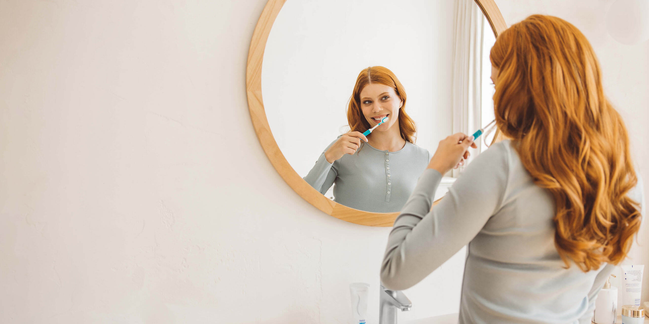 Eine Frau mit roten Haaren putzt sich die Zähne und betrachtet dabei ihr Spiegelbild in einem runden Badezimmerspiegel.