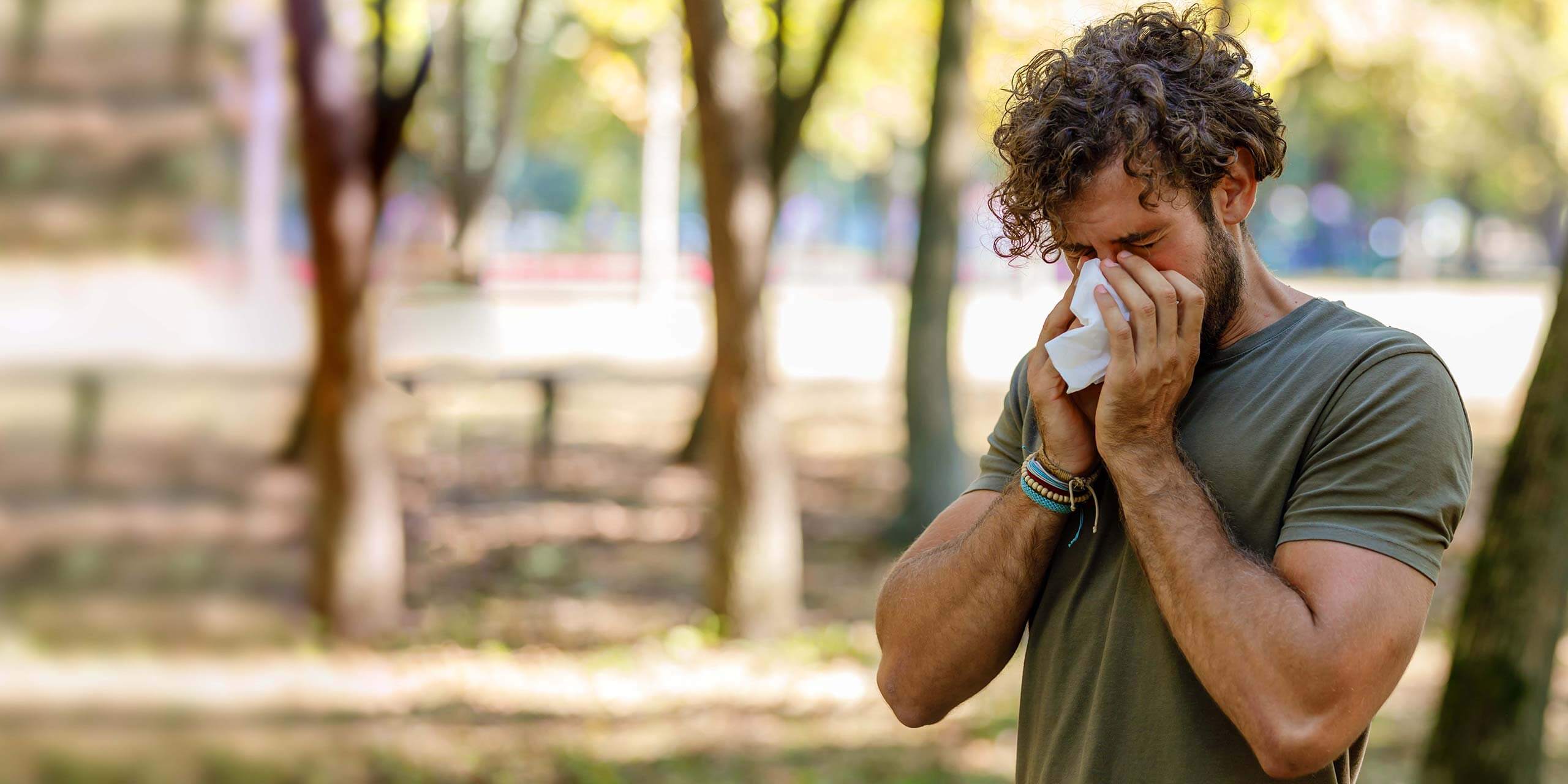 Ein Mann mit lockigem Haar steht draußen und putzt sich die Nase in ein Taschentuch, möglicherweise leidet er an Allergien oder einer Erkältung.