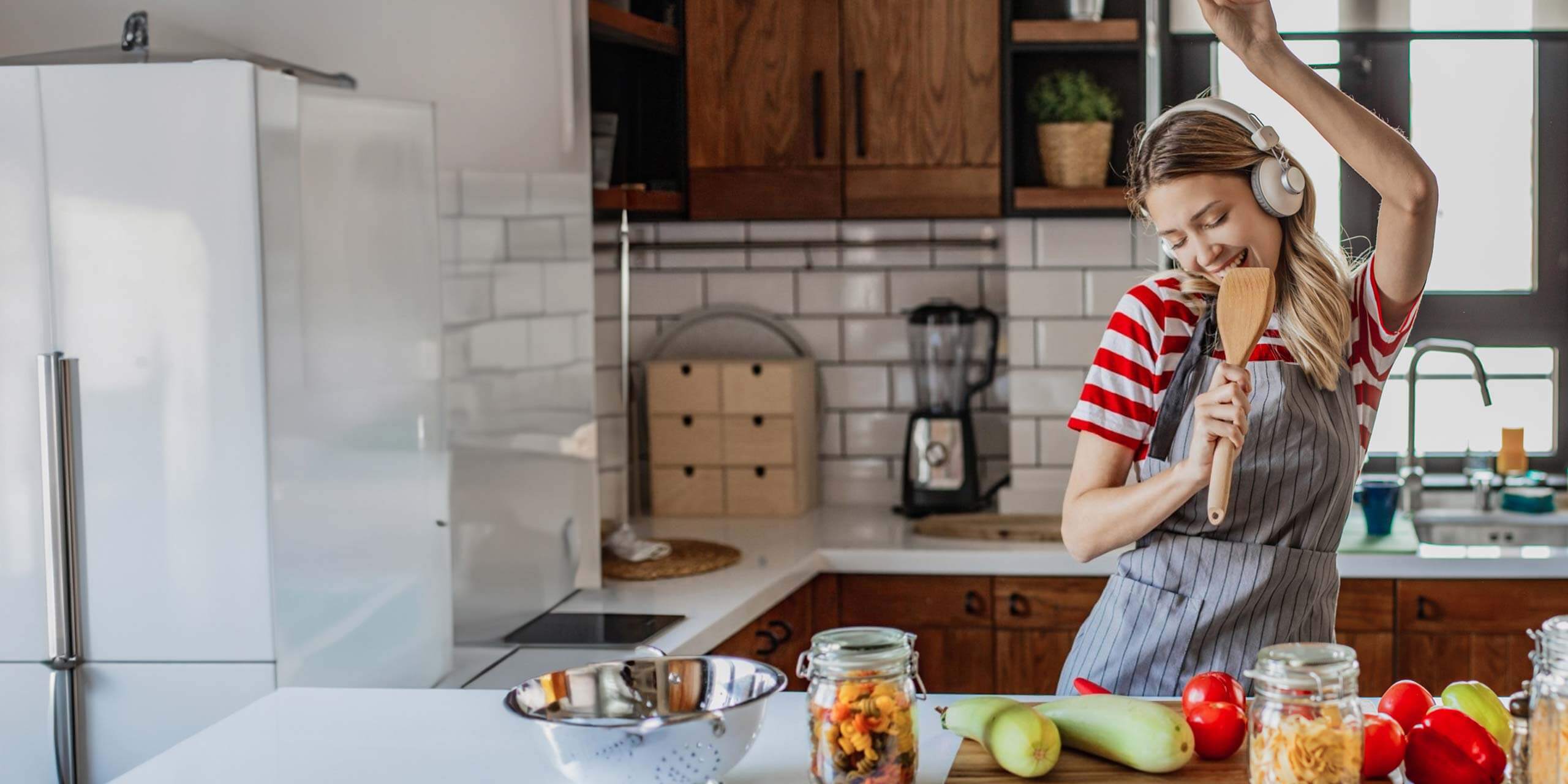 Eine fröhliche Person kocht in einer hellen Küche, tanzt, während sie einen Holzlöffel wie ein Mikrofon hält, während auf der Theke frische Zutaten darauf warten, zubereitet zu werden.